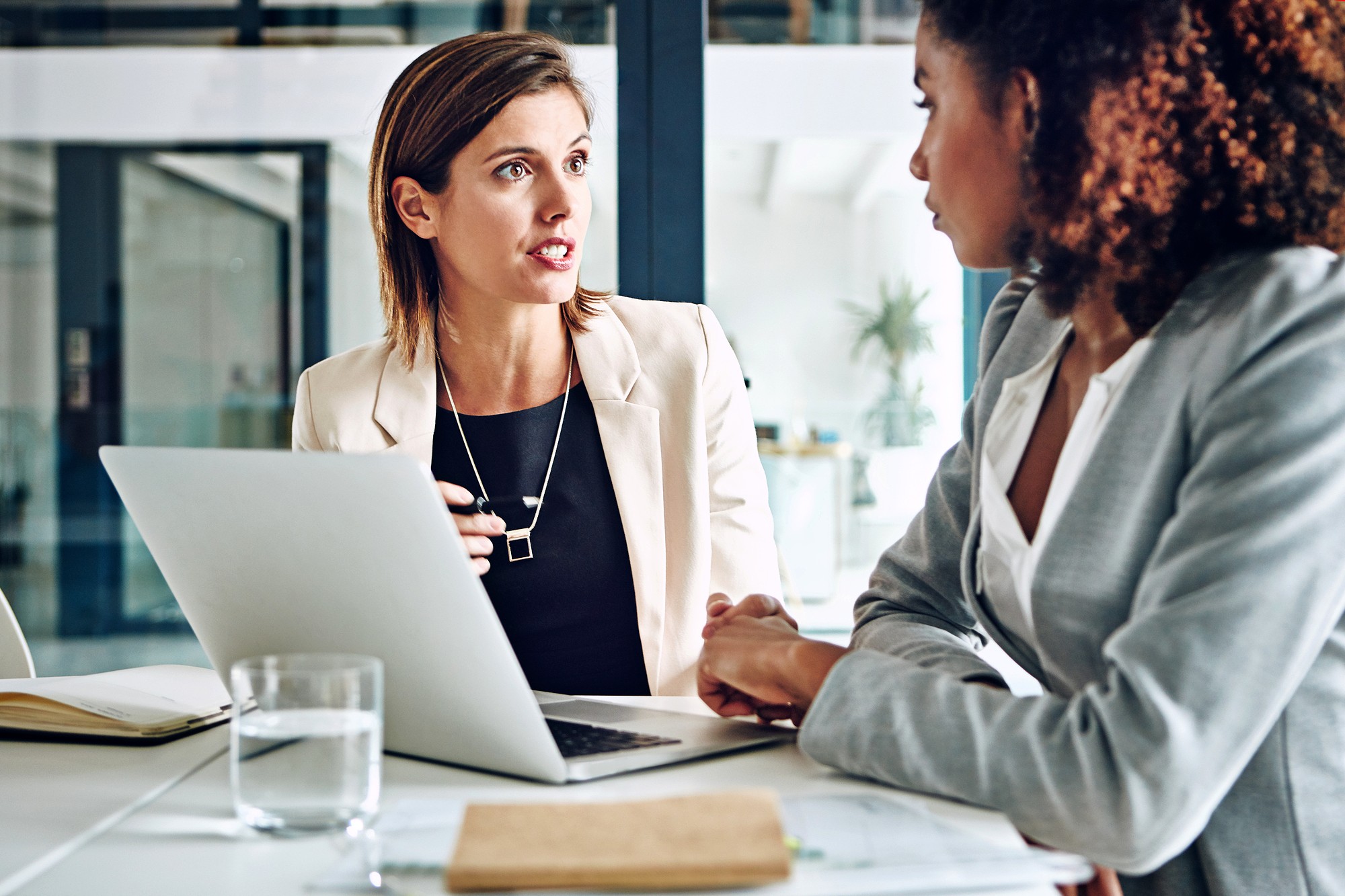 Two women in business attire sit at a desk in an office, engaged in a serious conversation. A laptop, notebook, and glass of water are on the desk in front of them.
