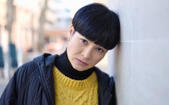 A person with short black hair and a serious expression leans their head against a light-colored wall. They are wearing a yellow knit sweater, a black turtleneck, and a dark jacket. The background is softly blurred.