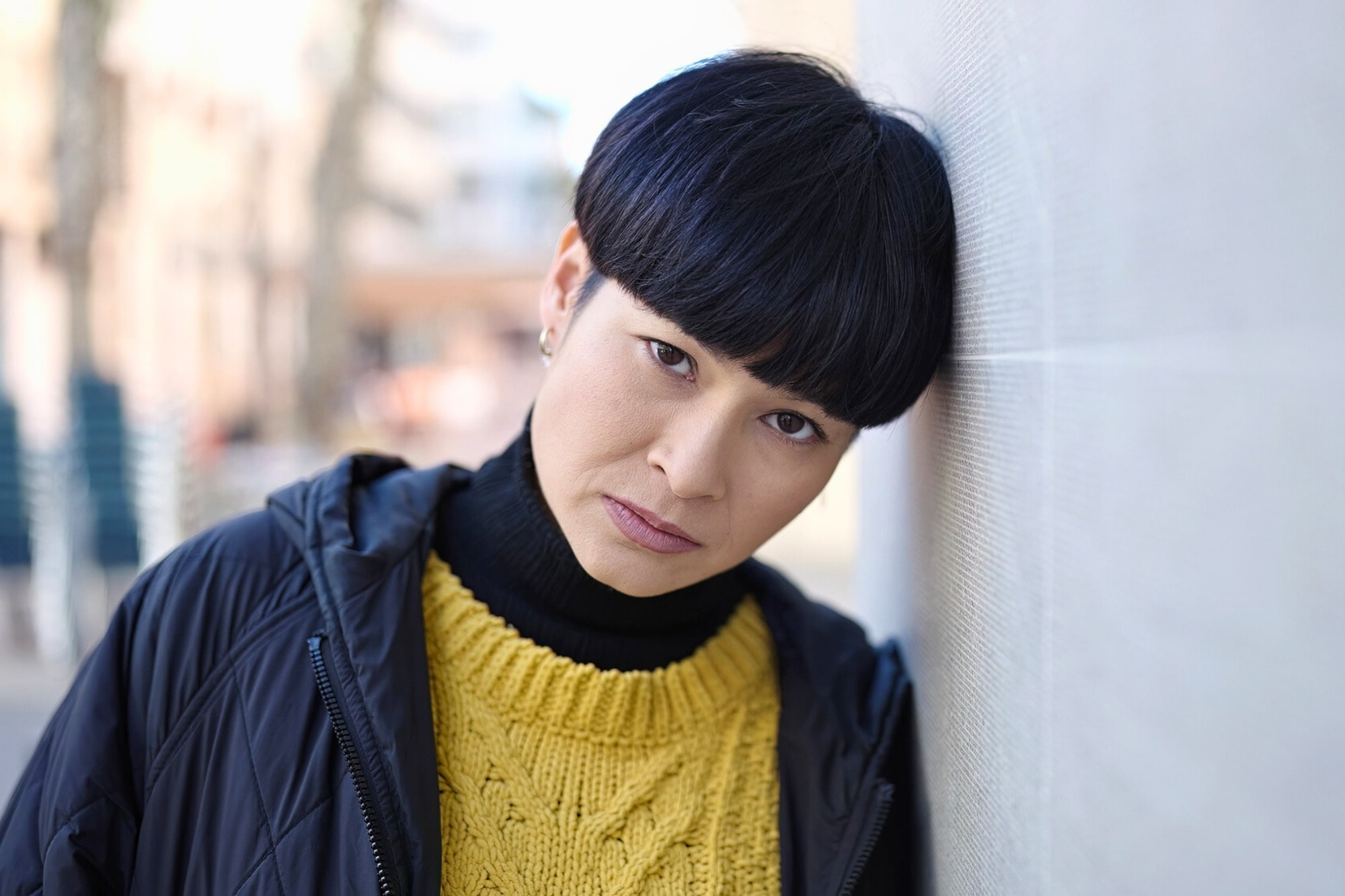 A person with short black hair and a serious expression leans their head against a light-colored wall. They are wearing a yellow knit sweater, a black turtleneck, and a dark jacket. The background is softly blurred.