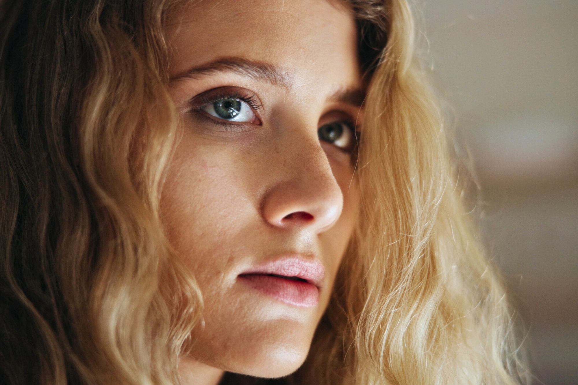 Close-up of a young woman with long, wavy blonde hair and blue eyes, looking slightly to the side with a neutral expression. The background is softly blurred.