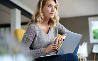 A woman with blonde hair sits on a yellow chair, holding a glass and resting her arm on an open laptop. She looks thoughtful and gazes into the distance in a modern, well-lit room.