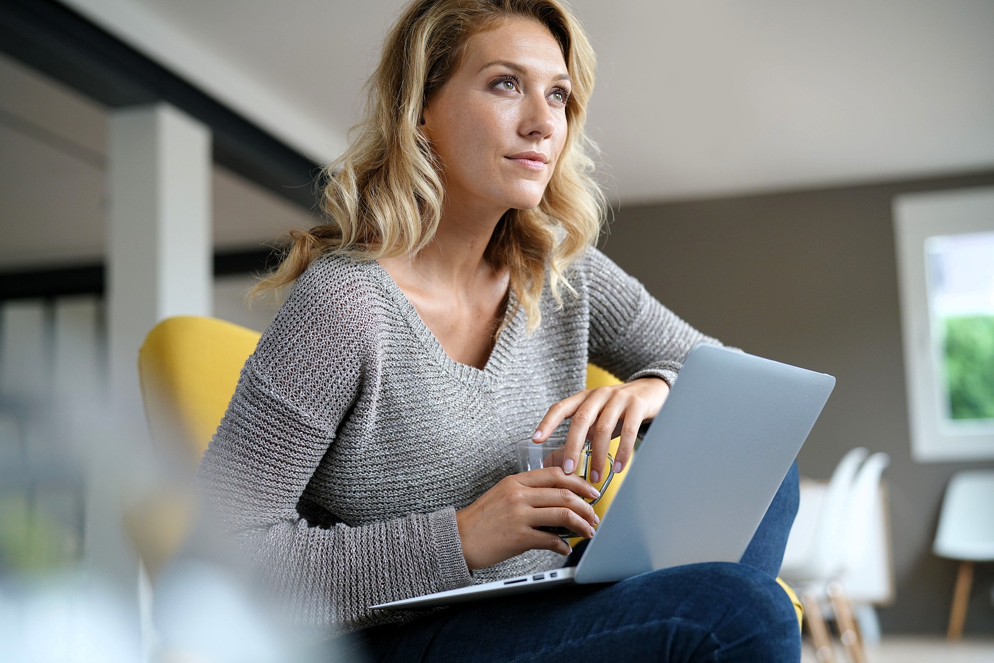 A woman with blonde hair sits on a yellow chair, holding a glass and resting her arm on an open laptop. She looks thoughtful and gazes into the distance in a modern, well-lit room.