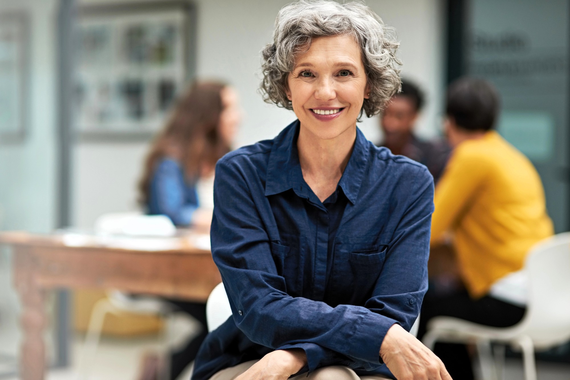 Smiling older woman with short curly gray hair wearing a blue shirt sits indoors with her arms crossed. In the blurred background, three people are talking around a table.