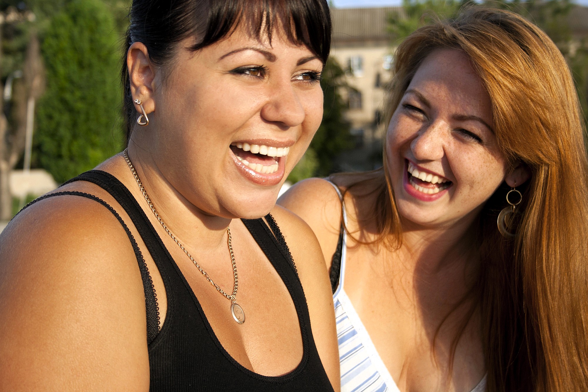 Two women are laughing together outdoors in bright sunlight. One has dark hair and wears a black tank top, while the other has long red hair and wears a striped top. Both appear happy and are enjoying each other's company.
