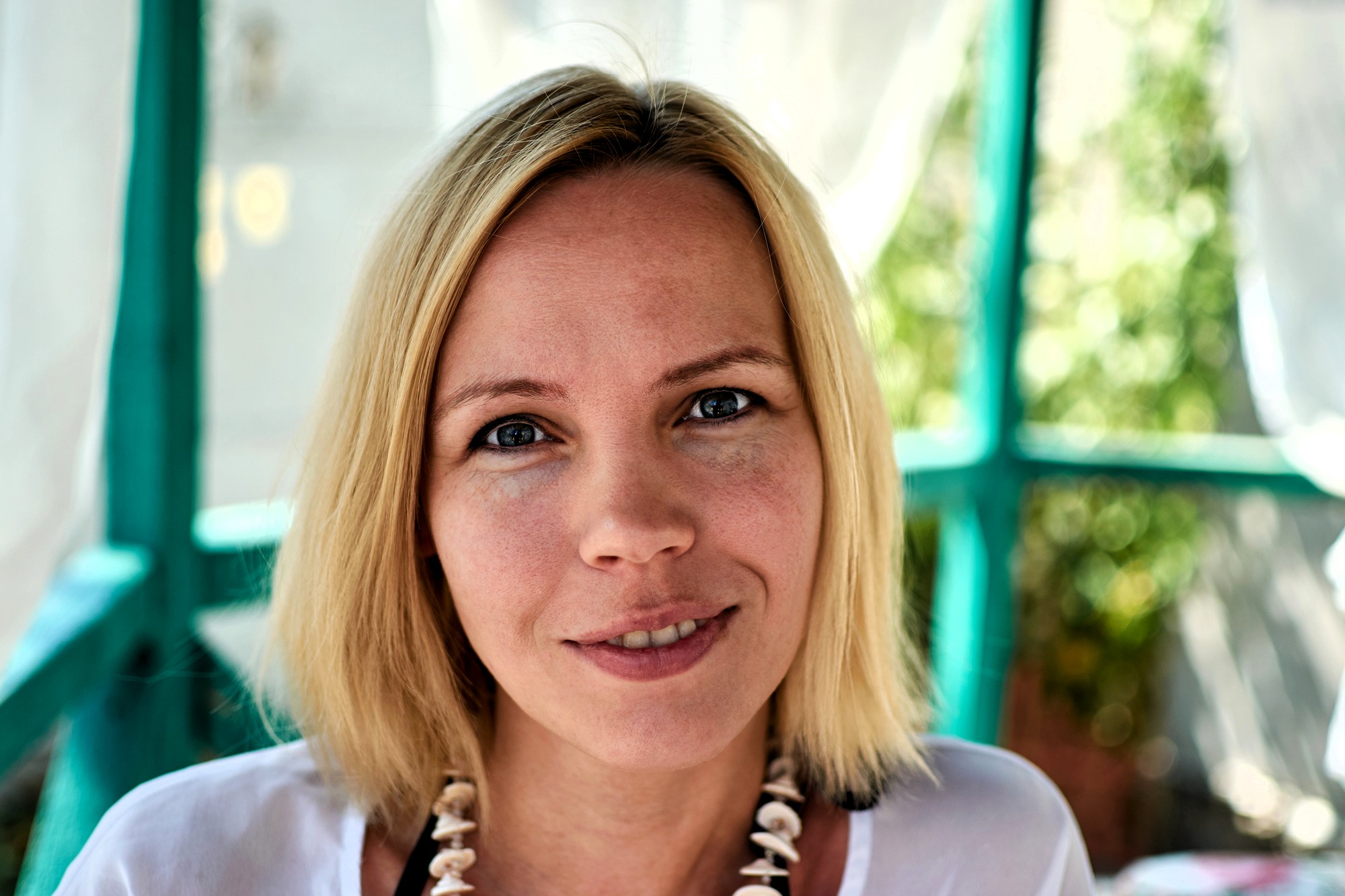 A woman with straight blonde hair and a shell necklace smiles softly at the camera. She is outdoors with green and white blurred structures and foliage in the background.