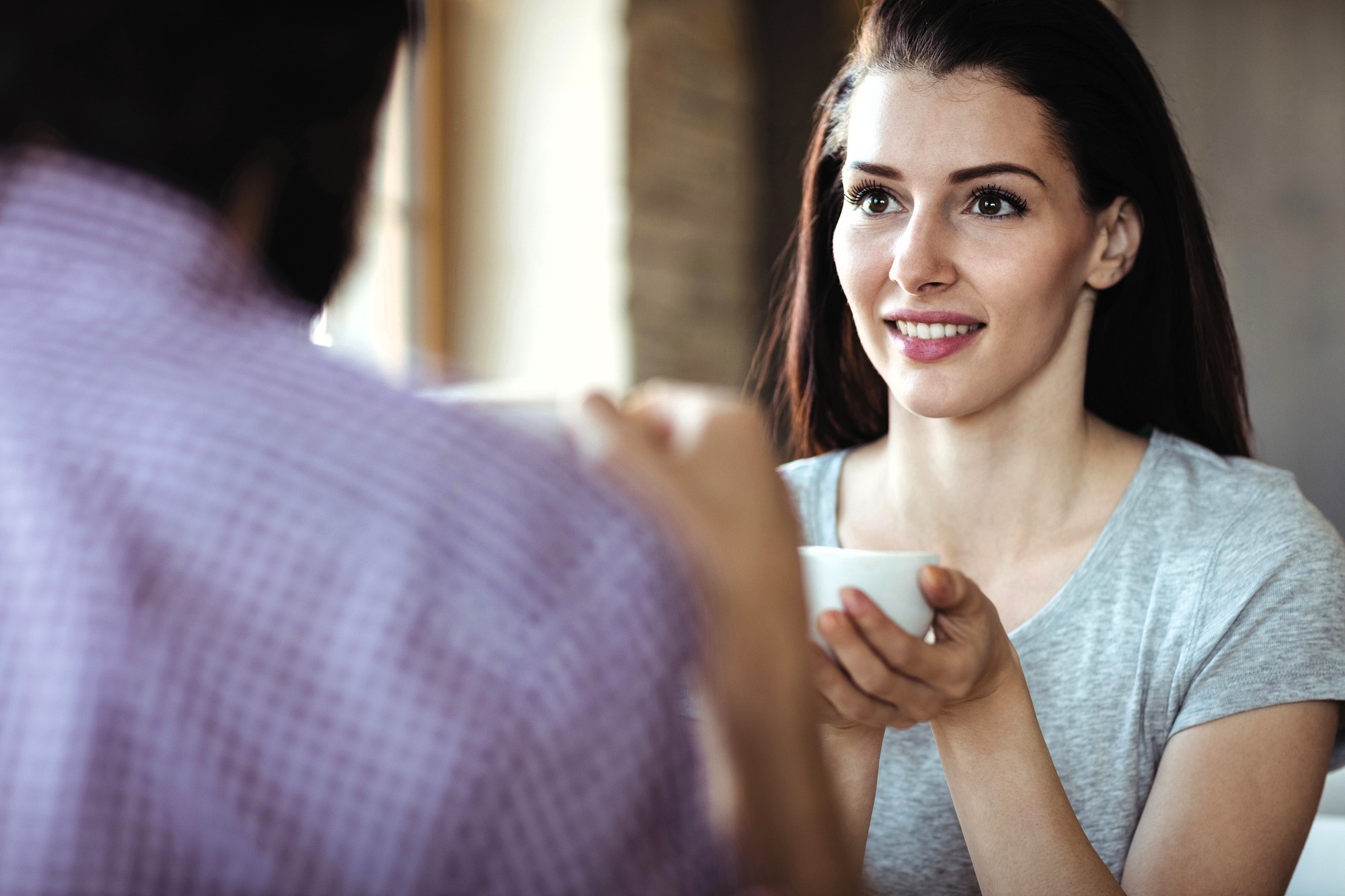 A woman with long brown hair smiles and holds a white coffee cup while sitting across from someone in a plaid shirt, engaging in conversation at a café.