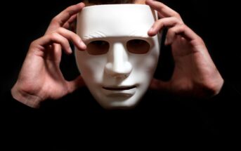 A person holds a white, expressionless mask in front of their face with both hands against a dark background, partially obscuring their features.