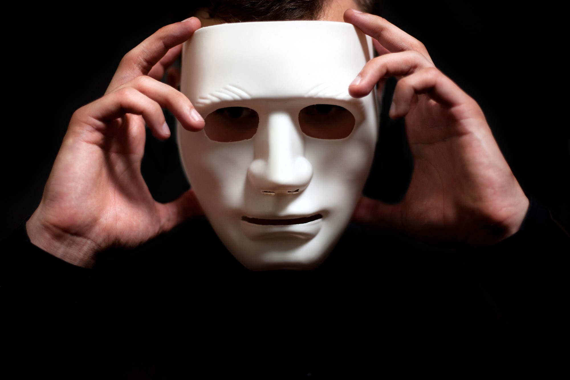 A person holds a white, expressionless mask in front of their face with both hands against a dark background, partially obscuring their features.