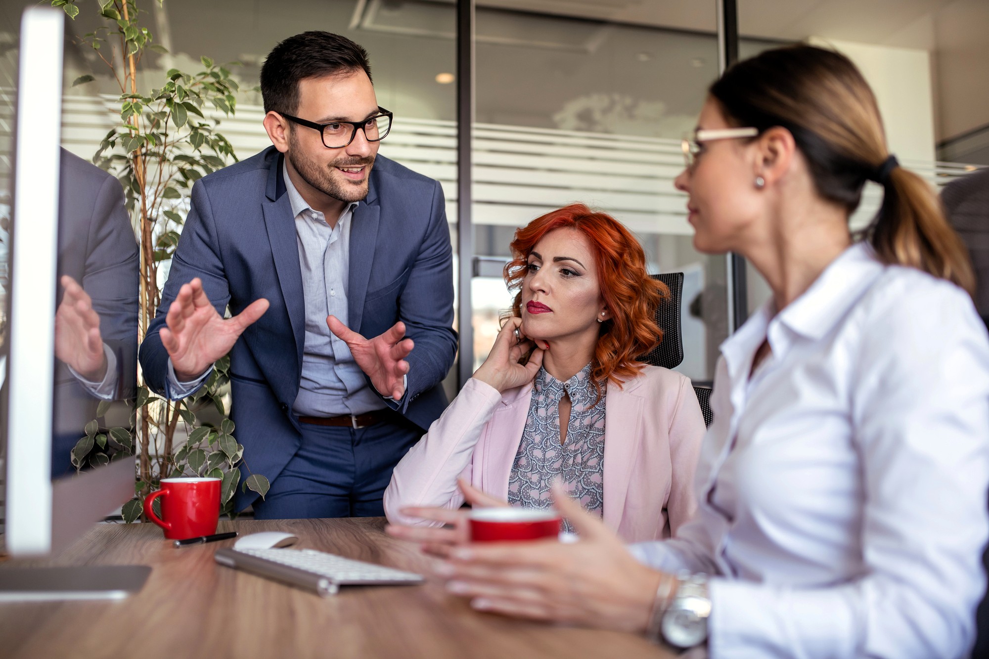 Three colleagues have a discussion at a desk in a modern office. A man in a suit gestures while talking to two women, one with red hair and the other with a ponytail, both listening attentively. Red coffee mugs are on the desk.