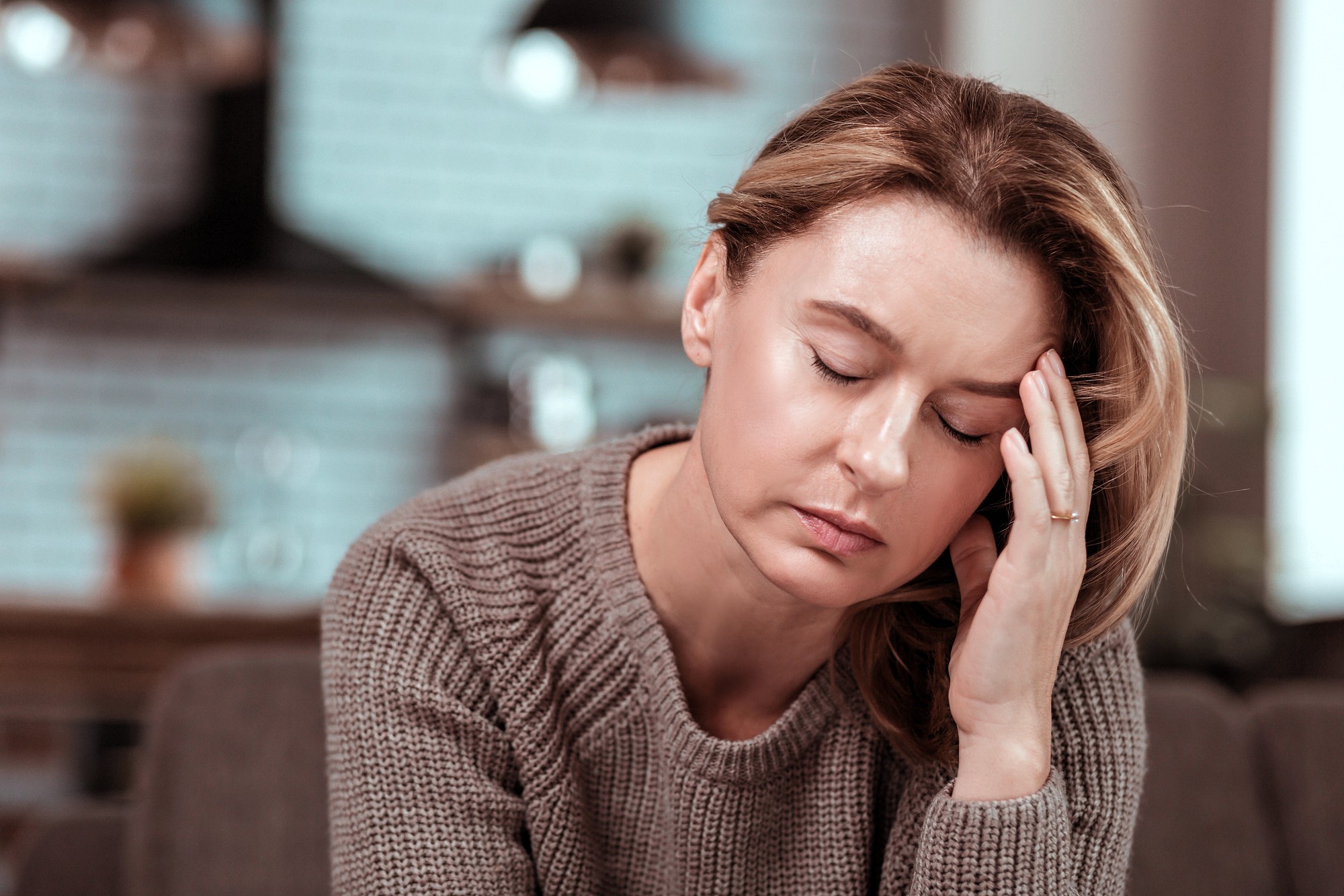 A woman with closed eyes rests her head on her hand, looking tired or stressed, wearing a knitted sweater indoors with a blurred background.