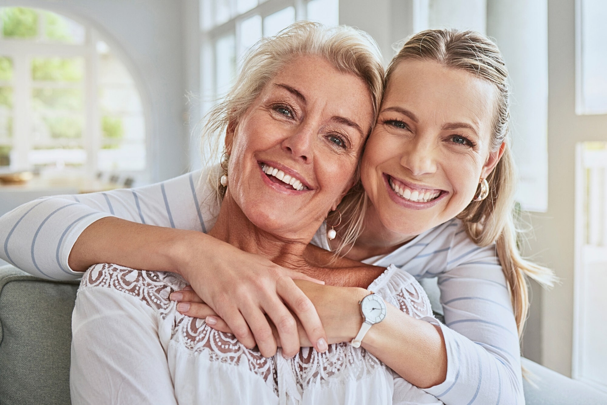 Two women sitting close together on a couch, smiling warmly at the camera. The younger woman hugs the older woman from behind. Both have light hair and are wearing light-colored, casual clothing in a bright, airy room.