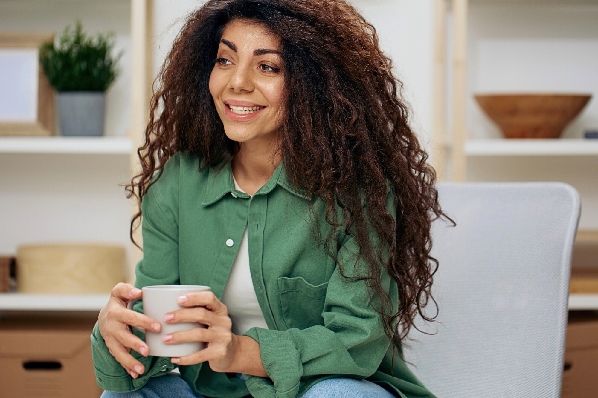 A young woman with long, curly hair sits on a chair, smiling and holding a mug. She is wearing a green shirt and jeans, with shelves and decorative items in the background.