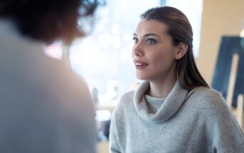 A woman with long brown hair wearing a light gray sweater looks attentively at someone out of focus in the foreground during a conversation in a bright indoor setting.