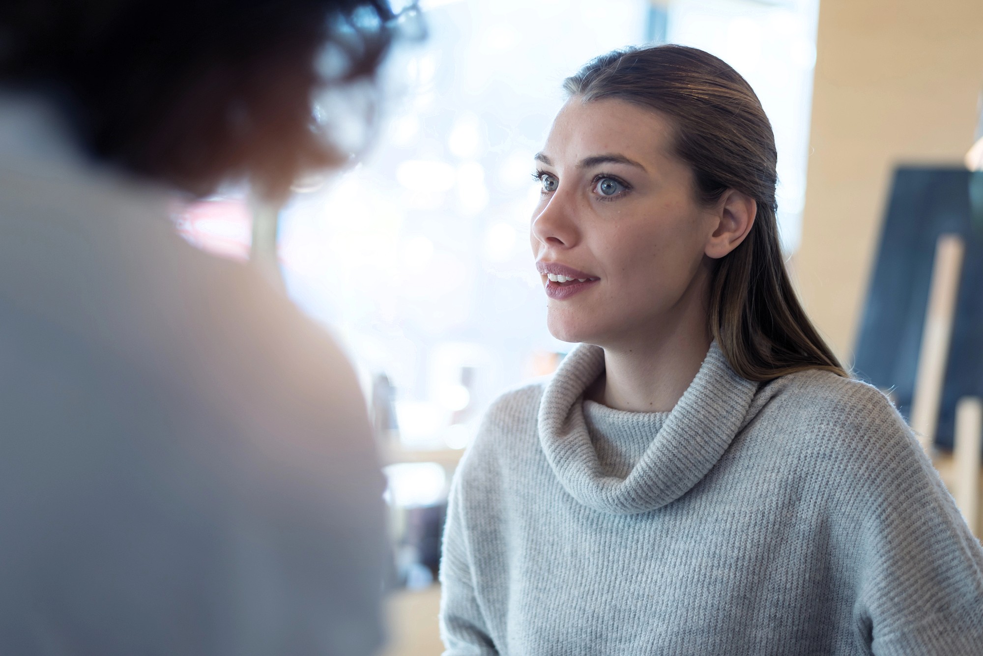 A woman with long brown hair wearing a light gray sweater looks attentively at someone out of focus in the foreground during a conversation in a bright indoor setting.
