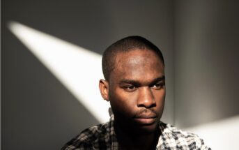 A man wearing a plaid shirt sits in soft sunlight, casting a triangle of light on the wall behind him. He looks down with a serious, contemplative expression, while shadows partially cover his face.