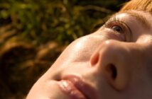 Close-up of a person's face lying down, with sunlight highlighting smooth skin, closed lips, and an eye gazing upward; background is blurred with green and brown tones.