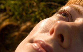 Close-up of a person's face lying down, with sunlight highlighting smooth skin, closed lips, and an eye gazing upward; background is blurred with green and brown tones.