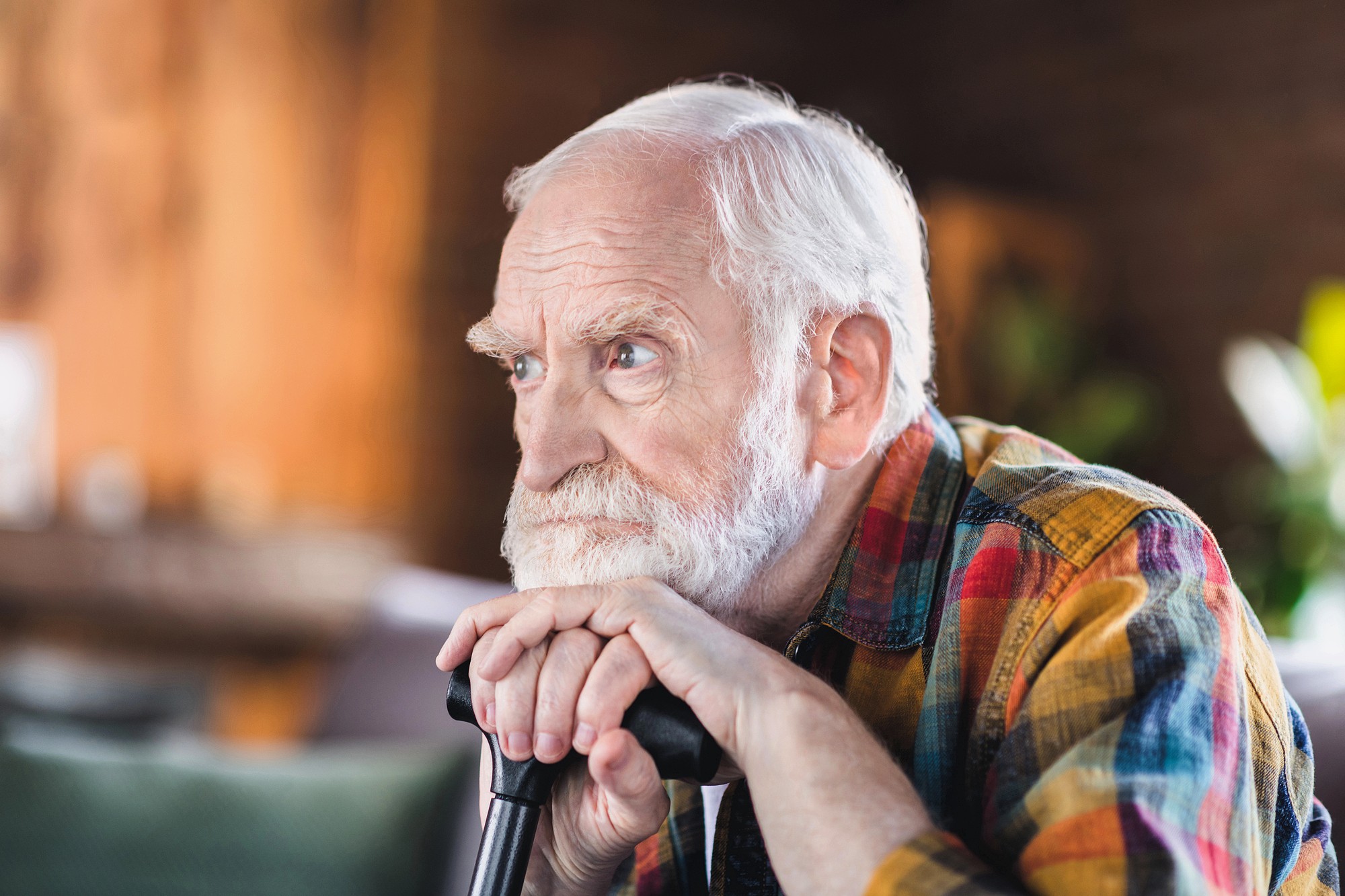 An elderly man with white hair and a beard is resting his hands and chin on a cane, looking thoughtfully into the distance. He is wearing a colorful plaid shirt and sitting indoors with a softly blurred background.