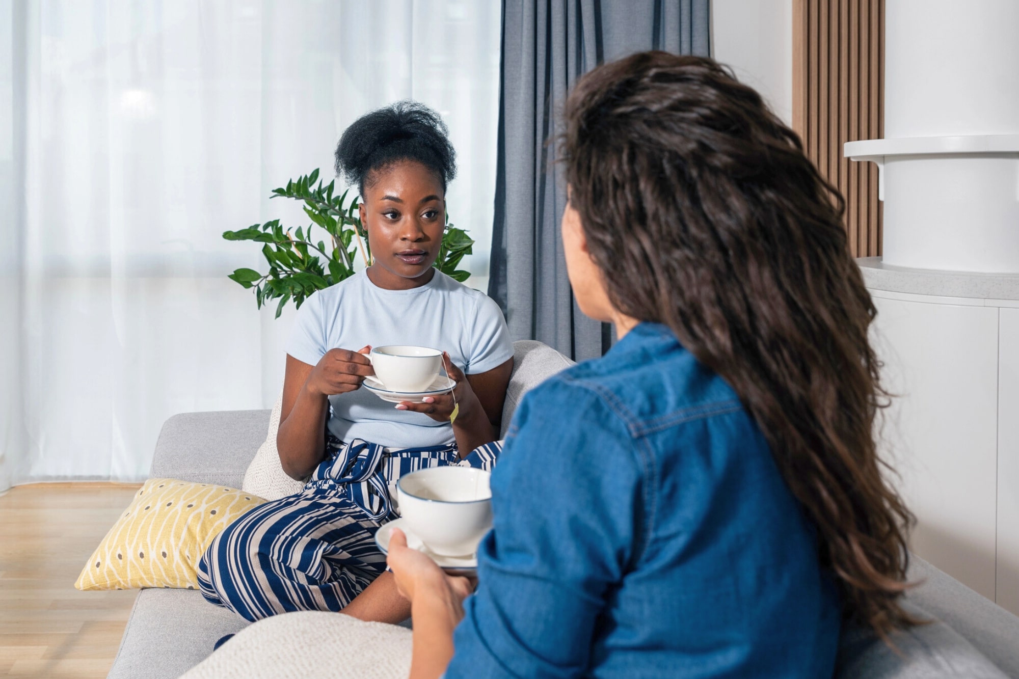 Two women sit on a couch indoors, holding cups and having a conversation. One woman faces the camera, while the other has her back to it. A plant and a window with sheer curtains are in the background.