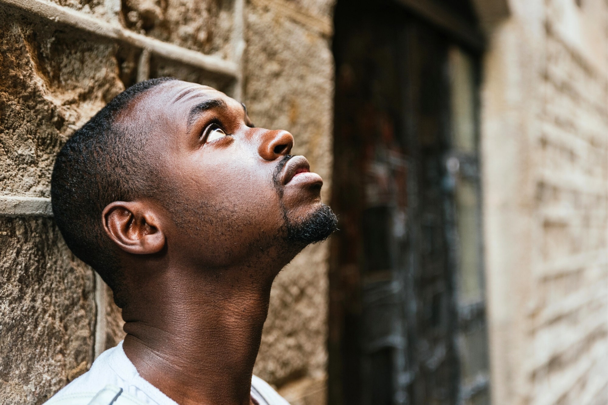 A young man with short hair and a trimmed beard stands next to a stone wall, looking upwards with a thoughtful expression. The background is slightly blurred, highlighting the texture of the wall.