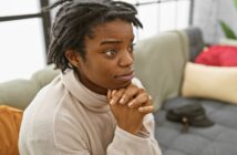 A person with short dreadlocks and a nose ring sits on a couch with their hands clasped under their chin, looking thoughtful or concerned. Pillows and a hat are visible in the background.