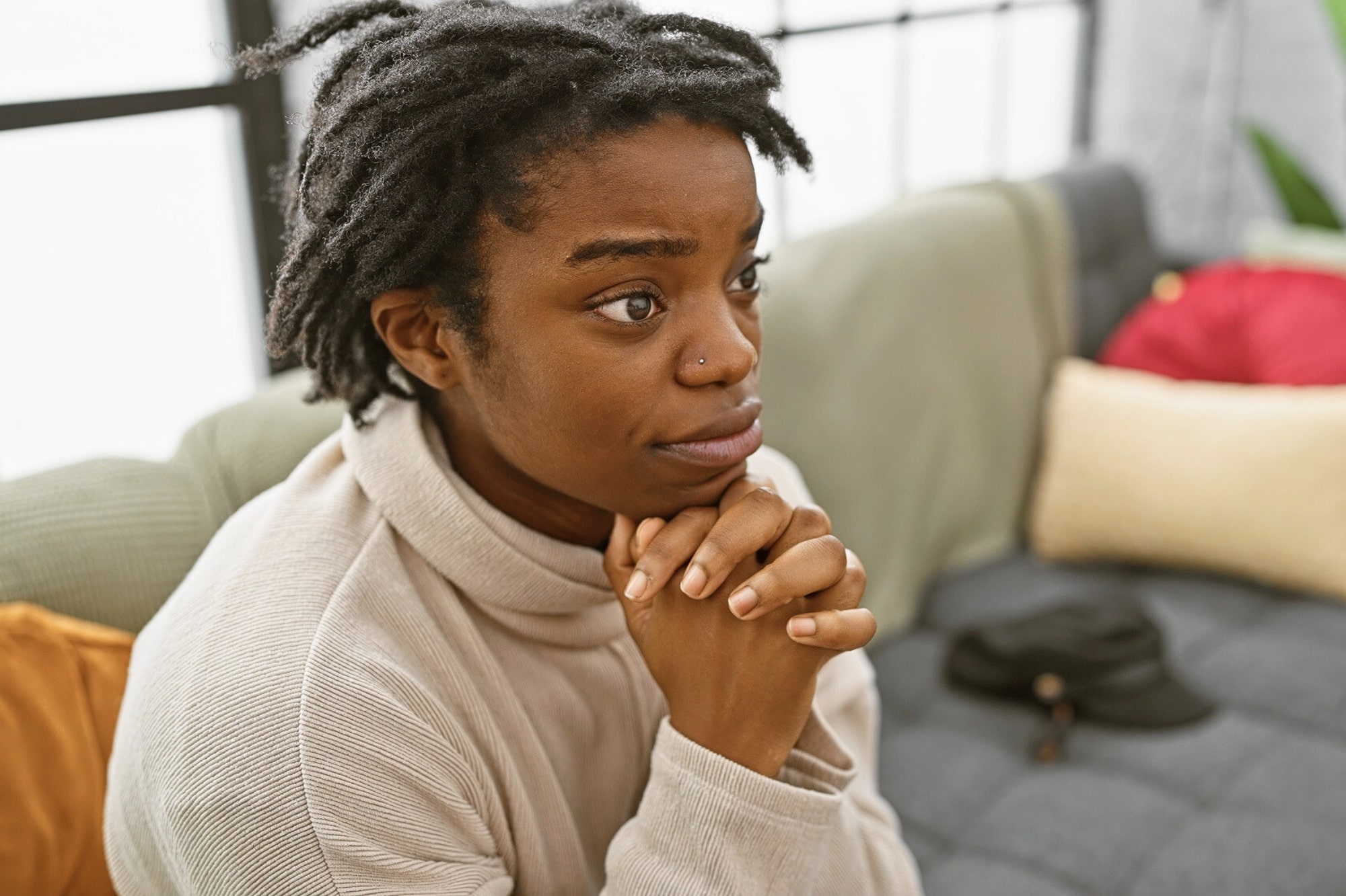 A person with short dreadlocks and a nose ring sits on a couch with their hands clasped under their chin, looking thoughtful or concerned. Pillows and a hat are visible in the background.