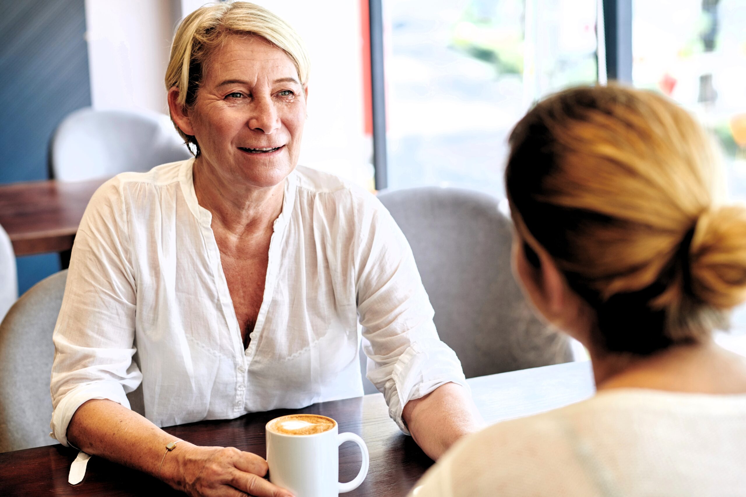 Two women sit across from each other at a cafe table, talking. One woman, with short blonde hair and a white shirt, holds a coffee mug and smiles warmly at the other, who has her back to the camera.