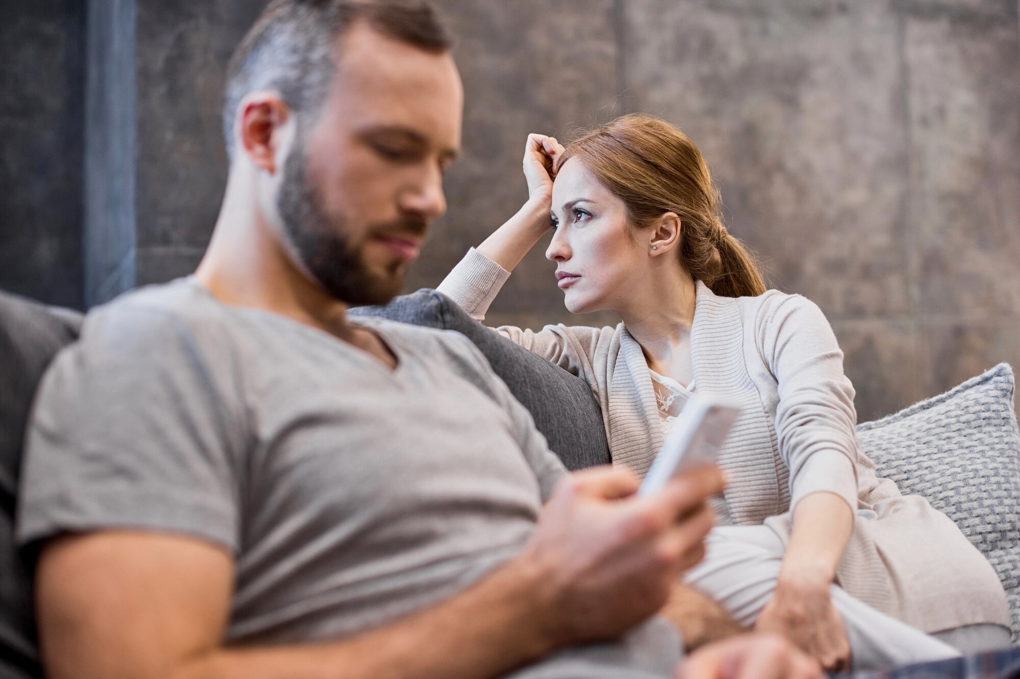 A woman sits on a couch looking concerned at a man who is focused on his phone, ignoring her. The atmosphere appears tense and distant between the two.