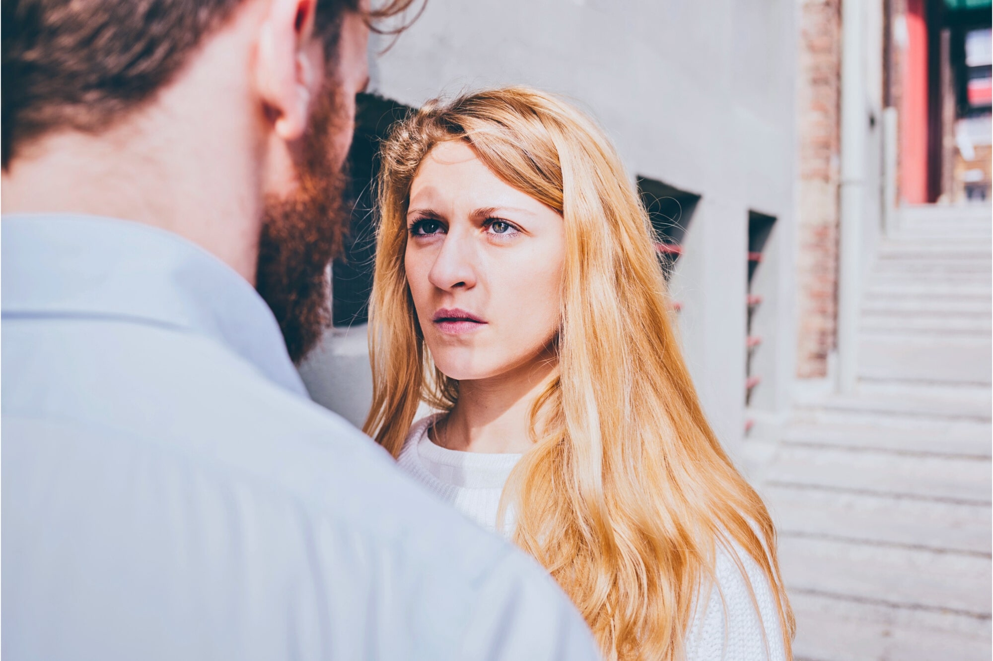 A woman with long blonde hair looks seriously at a man with a beard standing in front of her on an outdoor staircase, with sunlight illuminating her face.