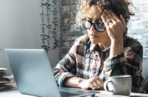 A woman wearing glasses and a plaid shirt sits at a desk with a laptop, holding her head in one hand and looking stressed. A coffee mug and paperwork are on the table next to her.