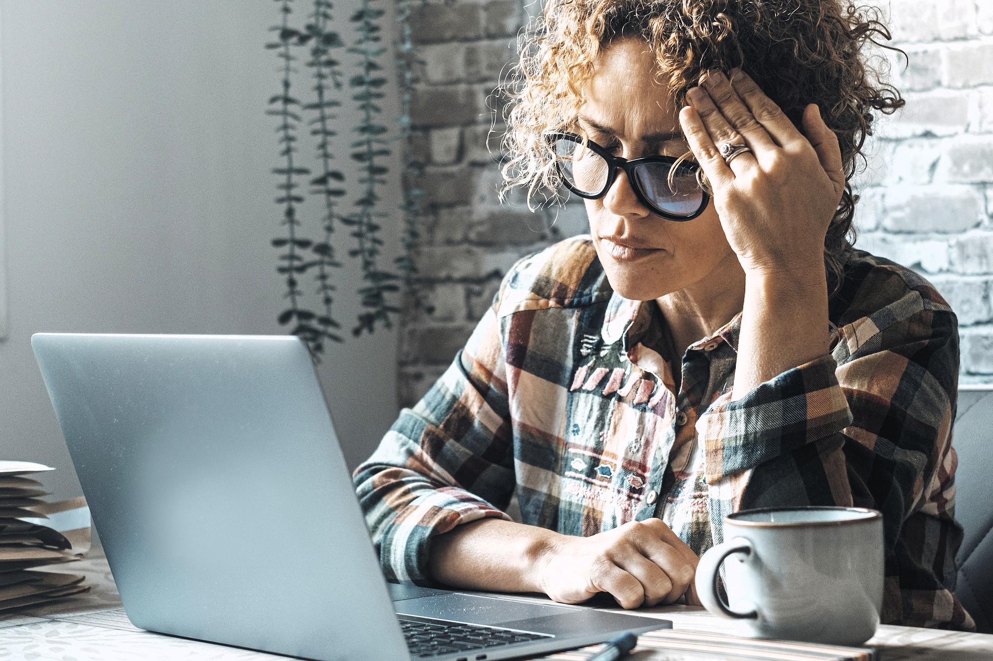 A woman wearing glasses and a plaid shirt sits at a desk with a laptop, holding her head in one hand and looking stressed. A coffee mug and paperwork are on the table next to her.