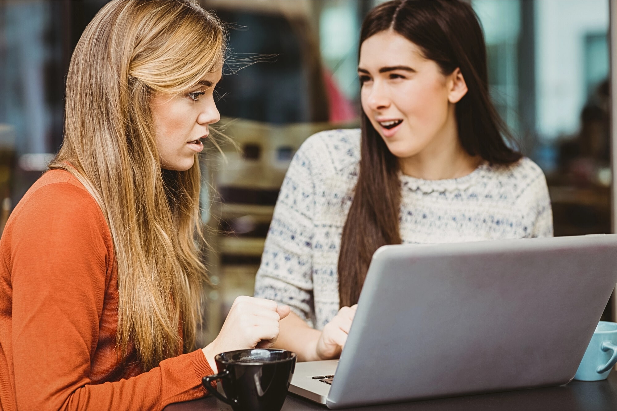 Two young women sit at a table with a laptop and coffee. One looks surprised or shocked while the other appears to explain something, both focusing on the laptop screen.