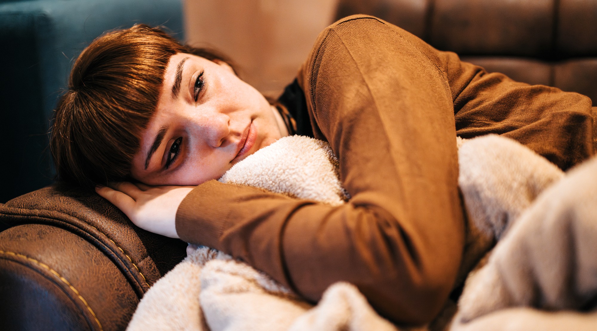 A woman with brown hair and bangs lies on a couch, resting her head on a pillow and hugging a soft beige blanket, looking tired or sad.