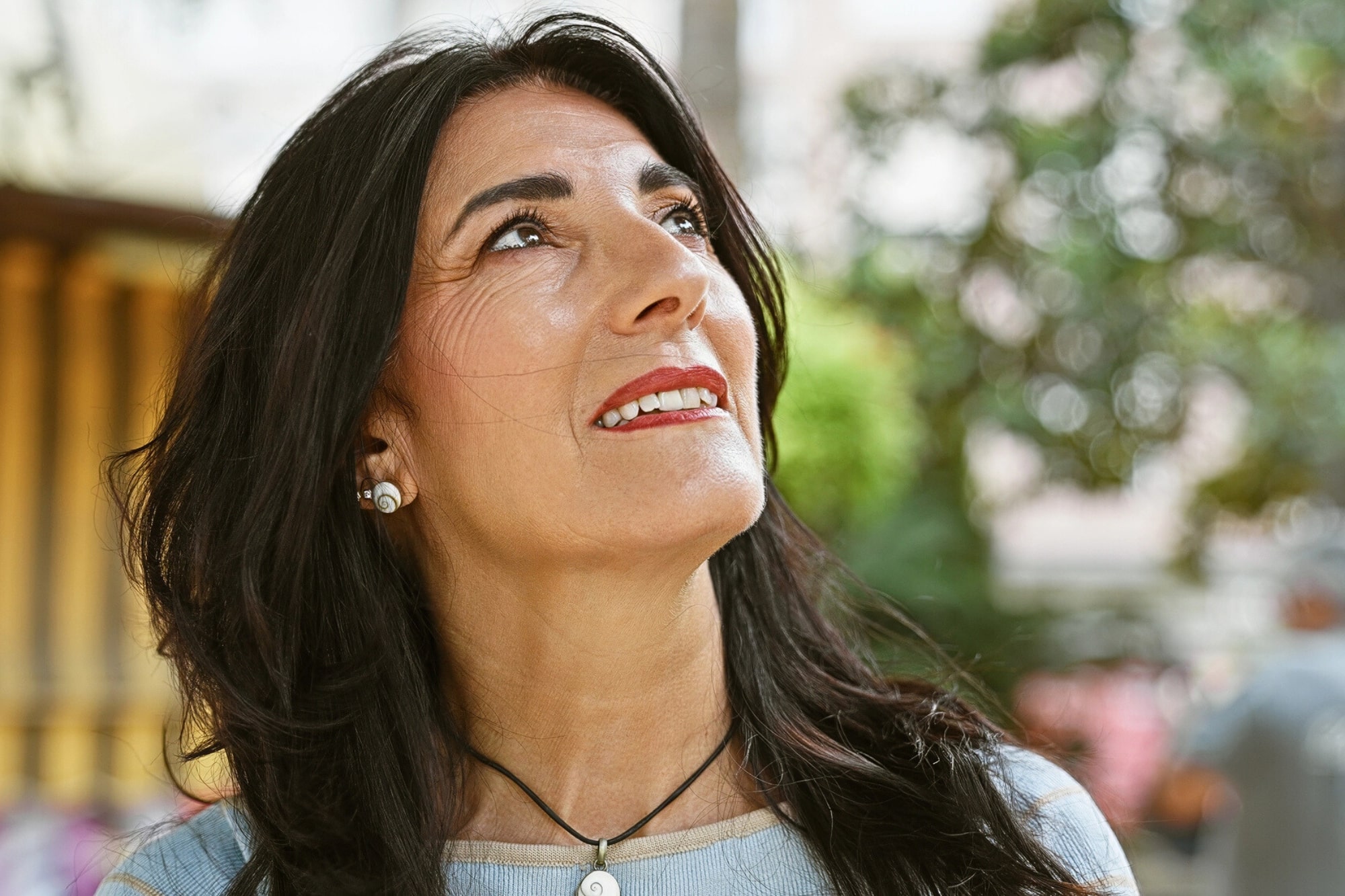 A woman with long dark hair and red lipstick looks up and smiles outdoors, with trees and blurred greenery in the background. She wears a light blue top, earrings, and a necklace.