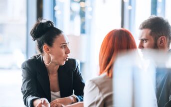 Three people are having a serious conversation at a table. A woman with dark hair in a bun and hoop earrings gestures while speaking to a man and a woman with red hair who are listening intently.
