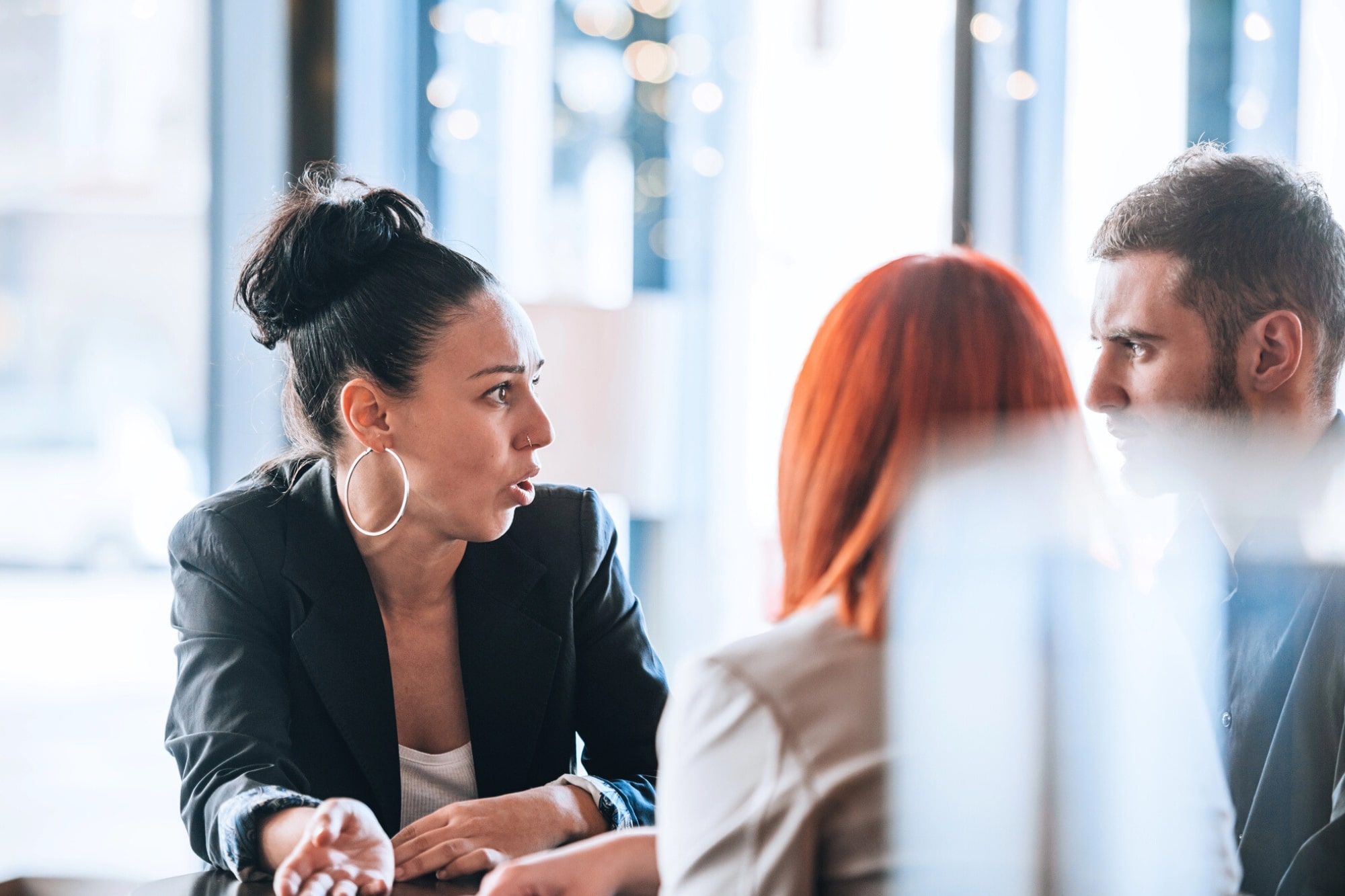 Three people are having a serious conversation at a table. A woman with dark hair in a bun and hoop earrings gestures while speaking to a man and a woman with red hair who are listening intently.
