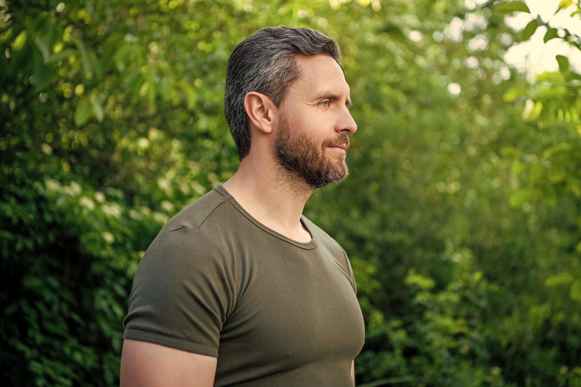 A man with short brown hair and a beard, wearing a green t-shirt, stands outdoors in front of lush green foliage, looking to the right with a calm expression.