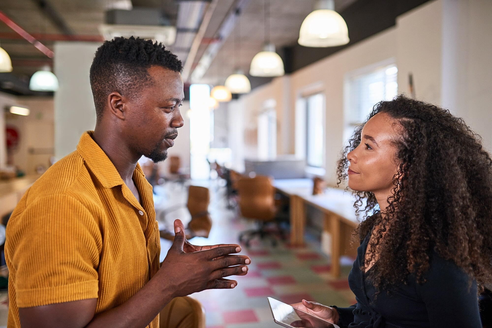 A man in a yellow shirt and a woman with curly hair are having a conversation in a modern, open-plan office. The woman is holding a smartphone and they appear engaged in discussion.