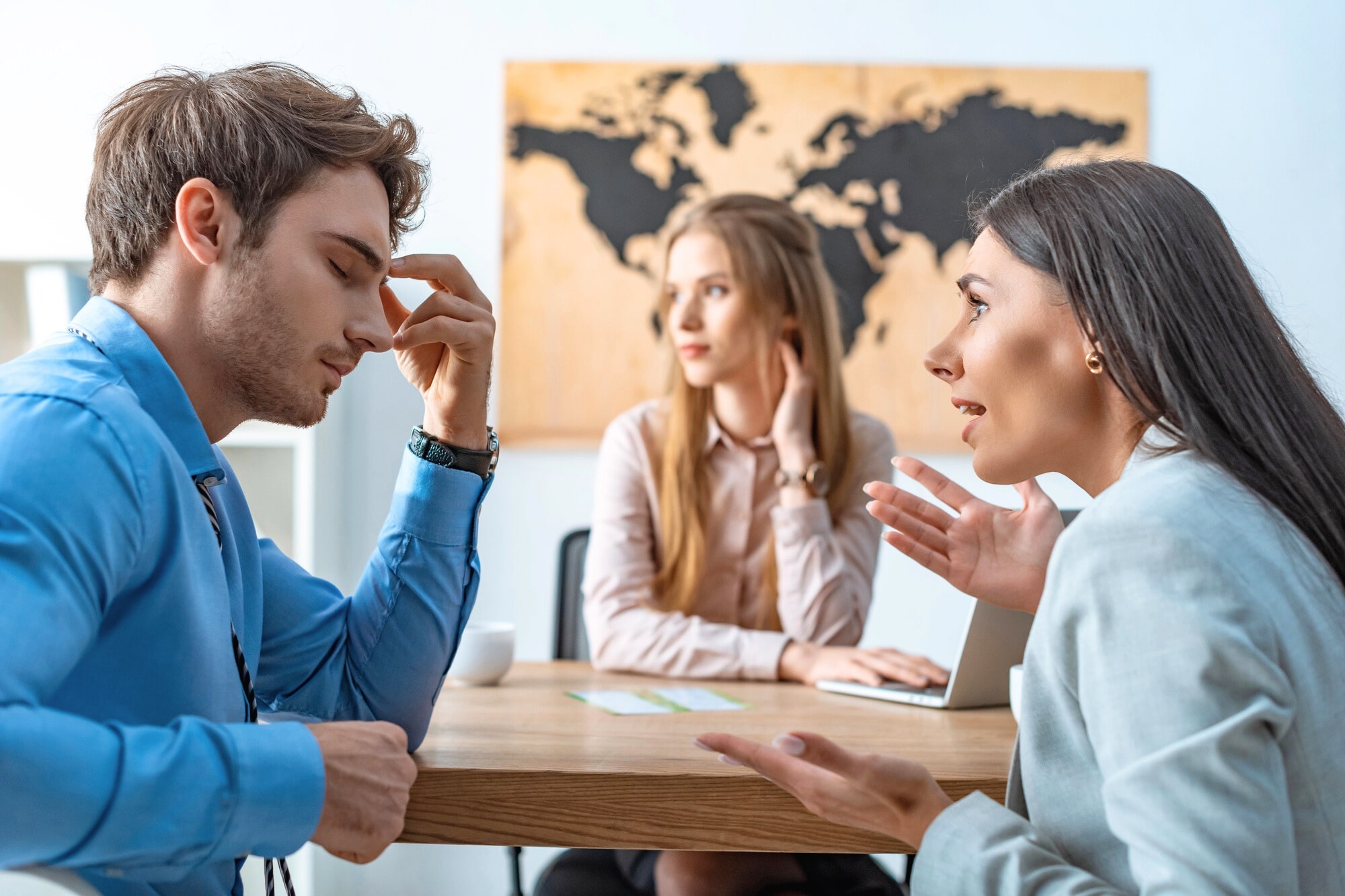 Three young professionals sit at a table in an office. One man looks stressed, touching his forehead; a woman gestures as she speaks to him; another woman in the background observes quietly. A world map is on the wall.