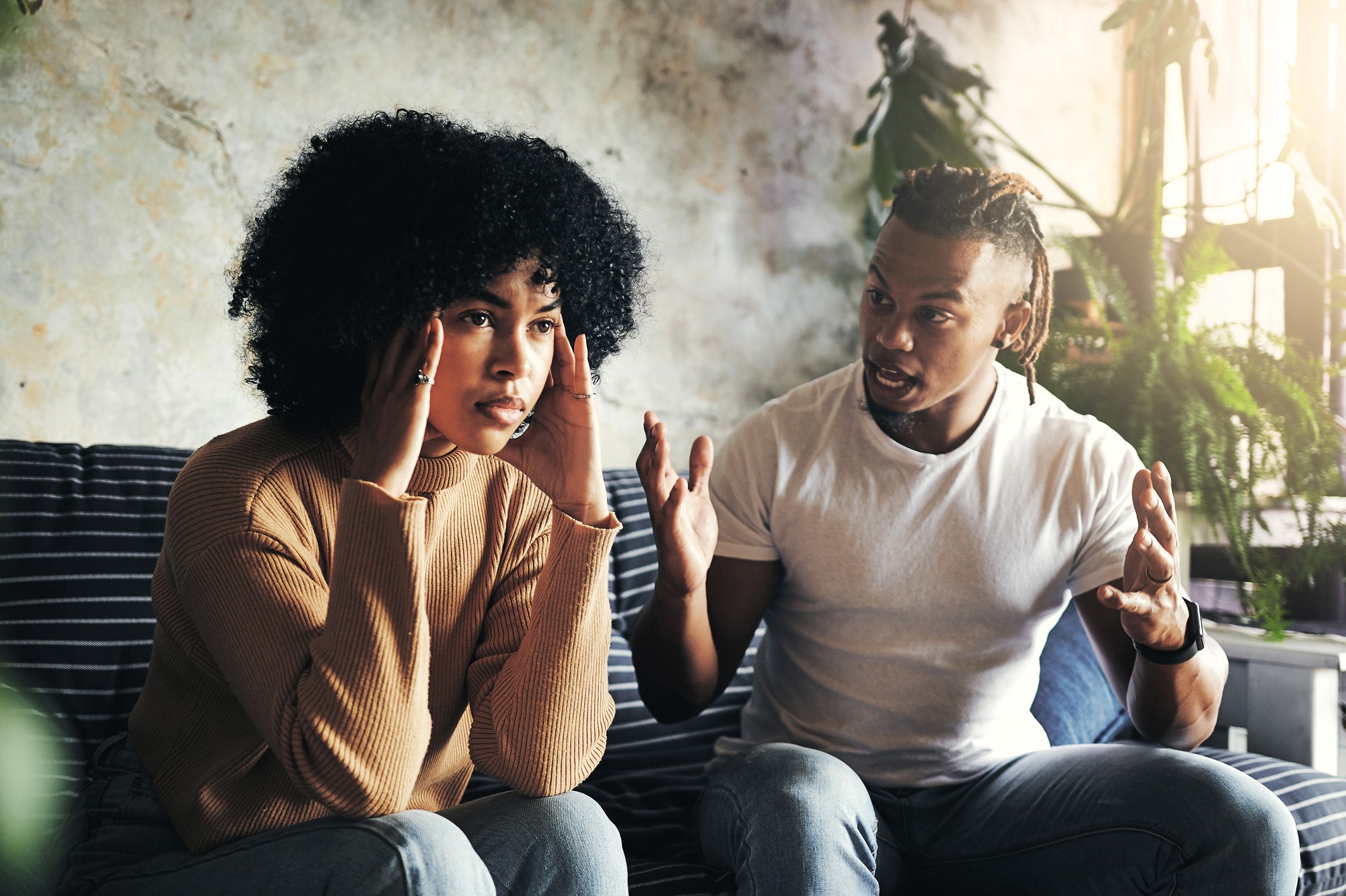 A woman sits on a couch touching her temples, looking frustrated, while a man next to her gestures with his hands and appears to be speaking intensely. They are indoors surrounded by plants.