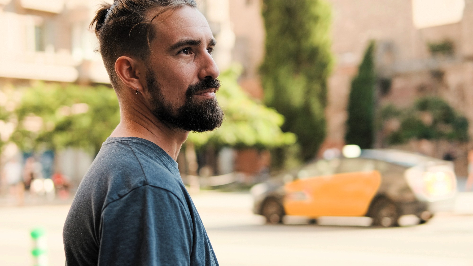 A bearded man with dark hair and a gray t-shirt stands outdoors, looking to the side. In the background, a yellow and black taxi passes by on a tree-lined city street.