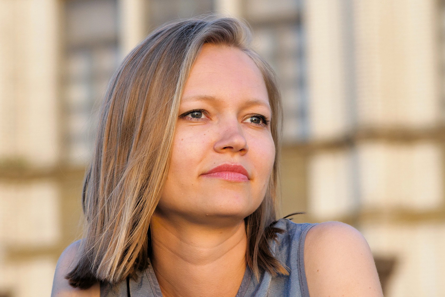 A woman with straight, shoulder-length blonde hair and blue eyes gazes thoughtfully into the distance, wearing a sleeveless gray top. The blurred background features a light-colored building.