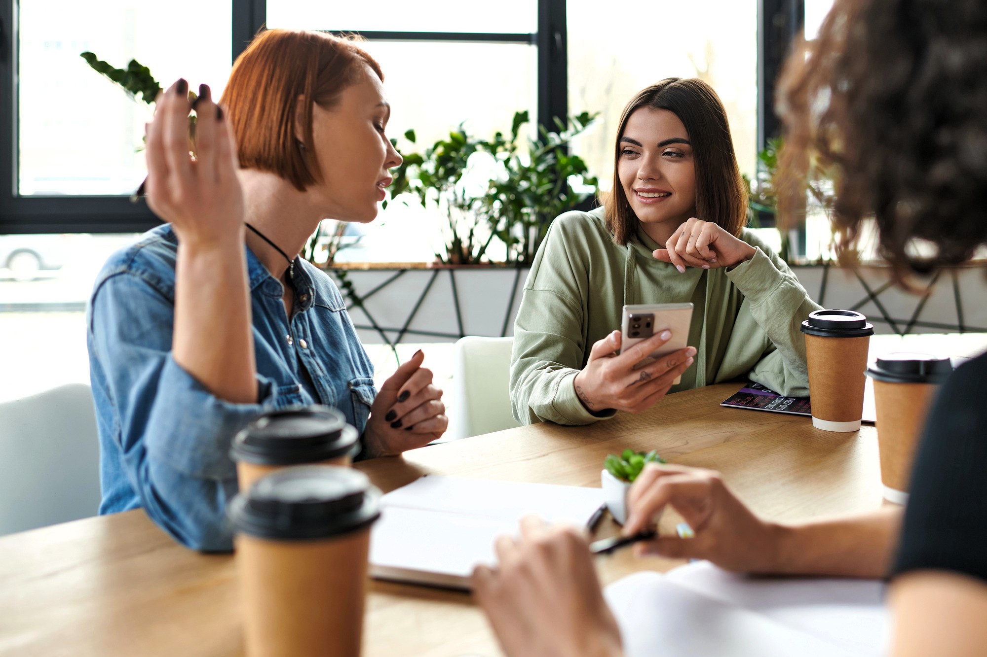 Three women sit at a table with notebooks and coffee cups, having a conversation. One woman holds a smartphone and smiles, while another gestures expressively. Sunlight streams through windows in the background.