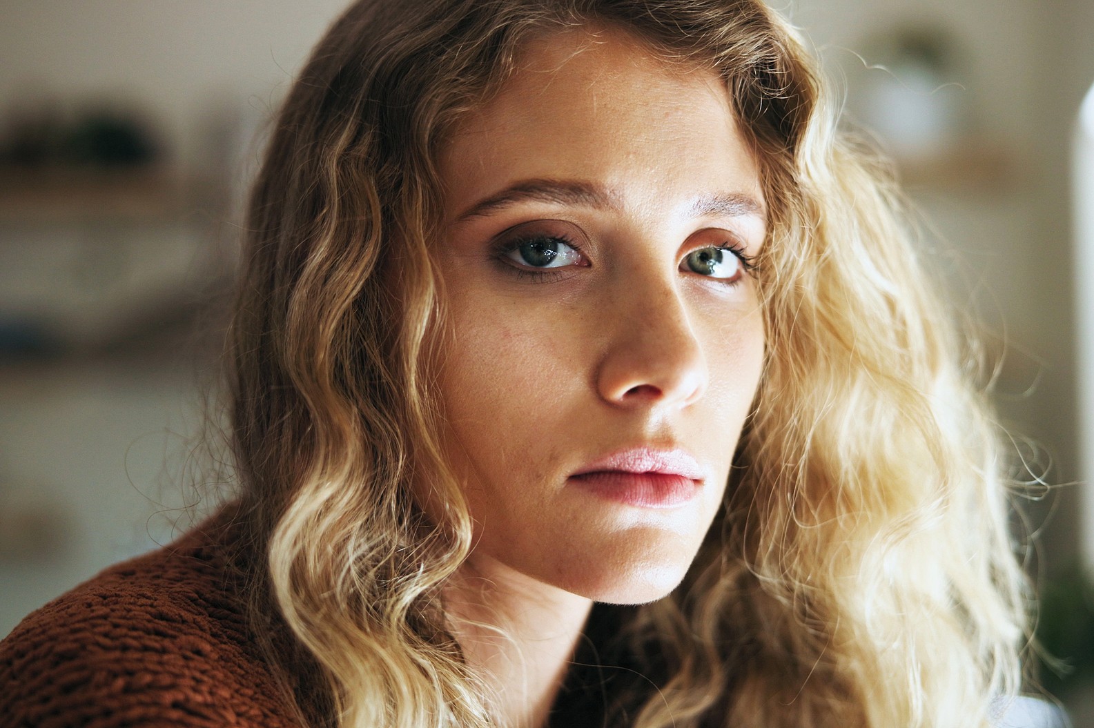 A woman with wavy blonde hair and blue eyes looks directly at the camera with a neutral expression. She is wearing a brown textured top, and the background is softly blurred.