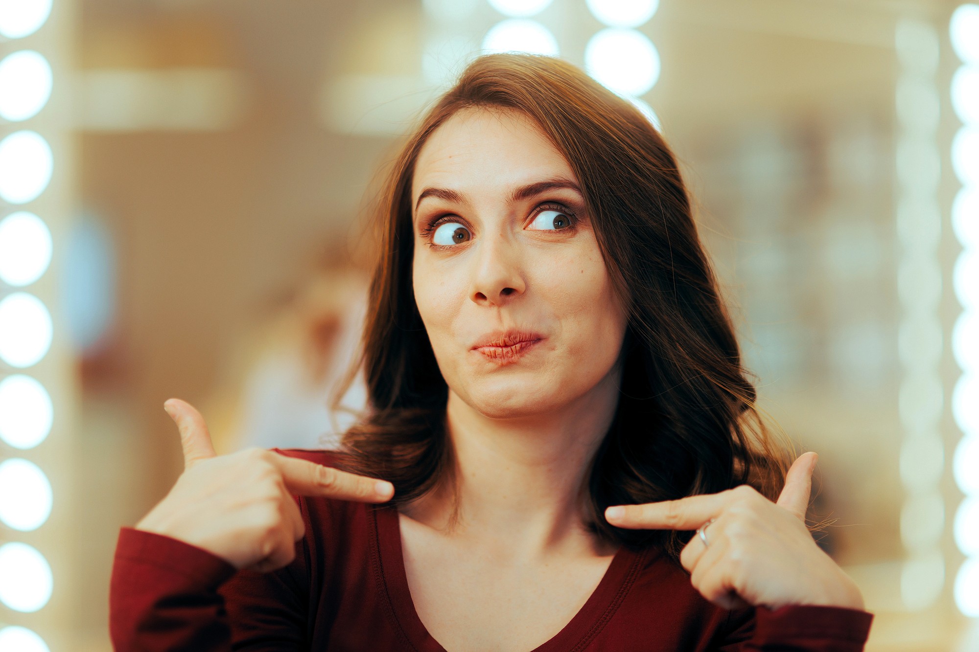 A woman with brown hair wearing a maroon top points at herself with both index fingers and makes a playful, surprised expression. The background is softly blurred with bright lights.