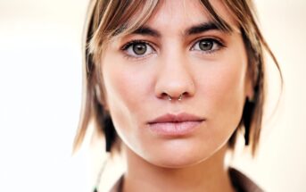 A young person with short brown hair and bangs, wearing a double nose ring and minimal makeup, looks directly at the camera with a neutral expression. The background is softly blurred.