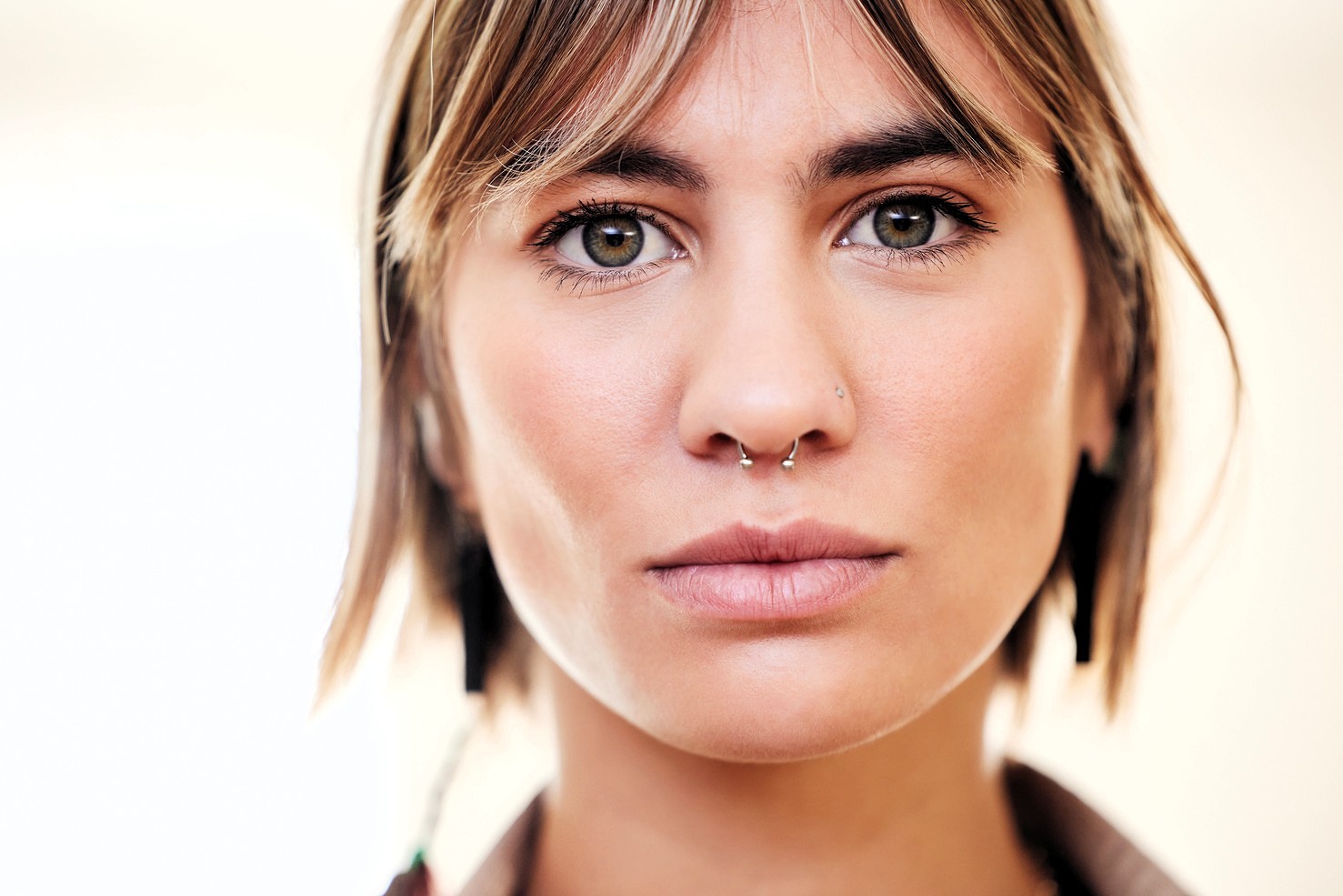 A young person with short brown hair and bangs, wearing a double nose ring and minimal makeup, looks directly at the camera with a neutral expression. The background is softly blurred.