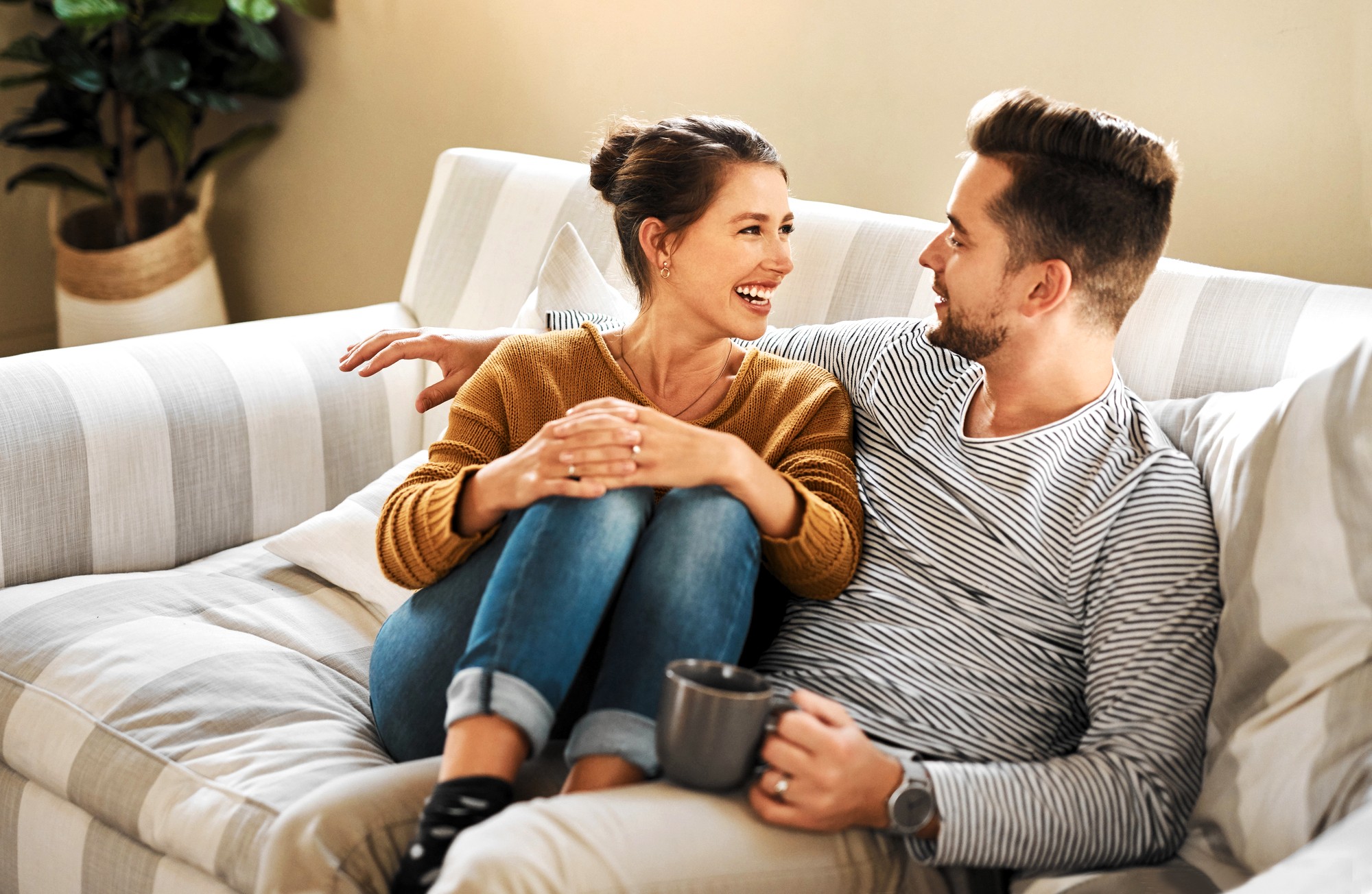 A woman and a man sit closely together on a striped couch, smiling and looking at each other. The woman wears a brown sweater and jeans, while the man holds a mug and wears a striped shirt. A plant is visible in the background.