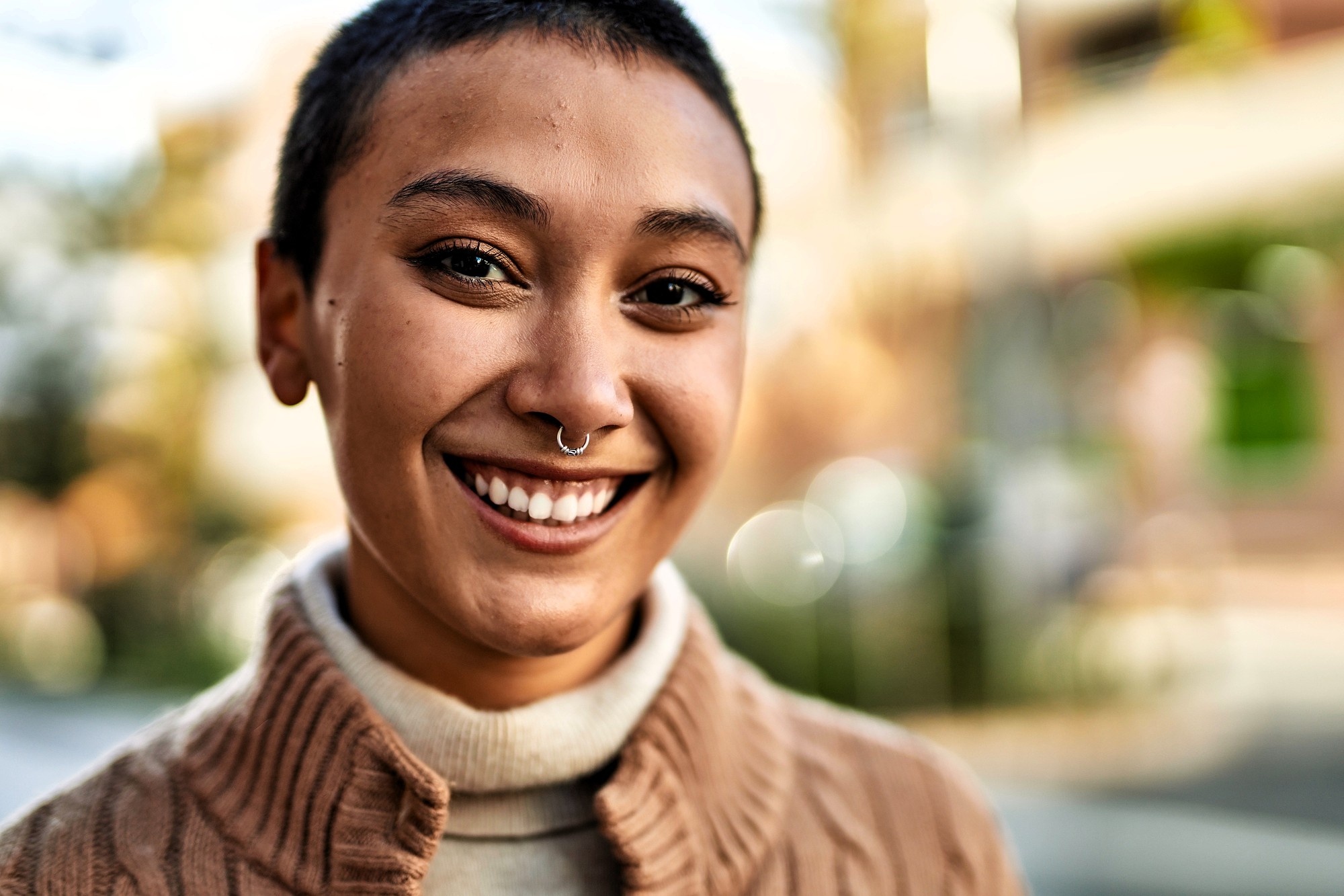 A person with short hair and a nose ring smiles outdoors, wearing a beige turtleneck and a brown textured sweater. The background is blurred with natural light and soft colors.