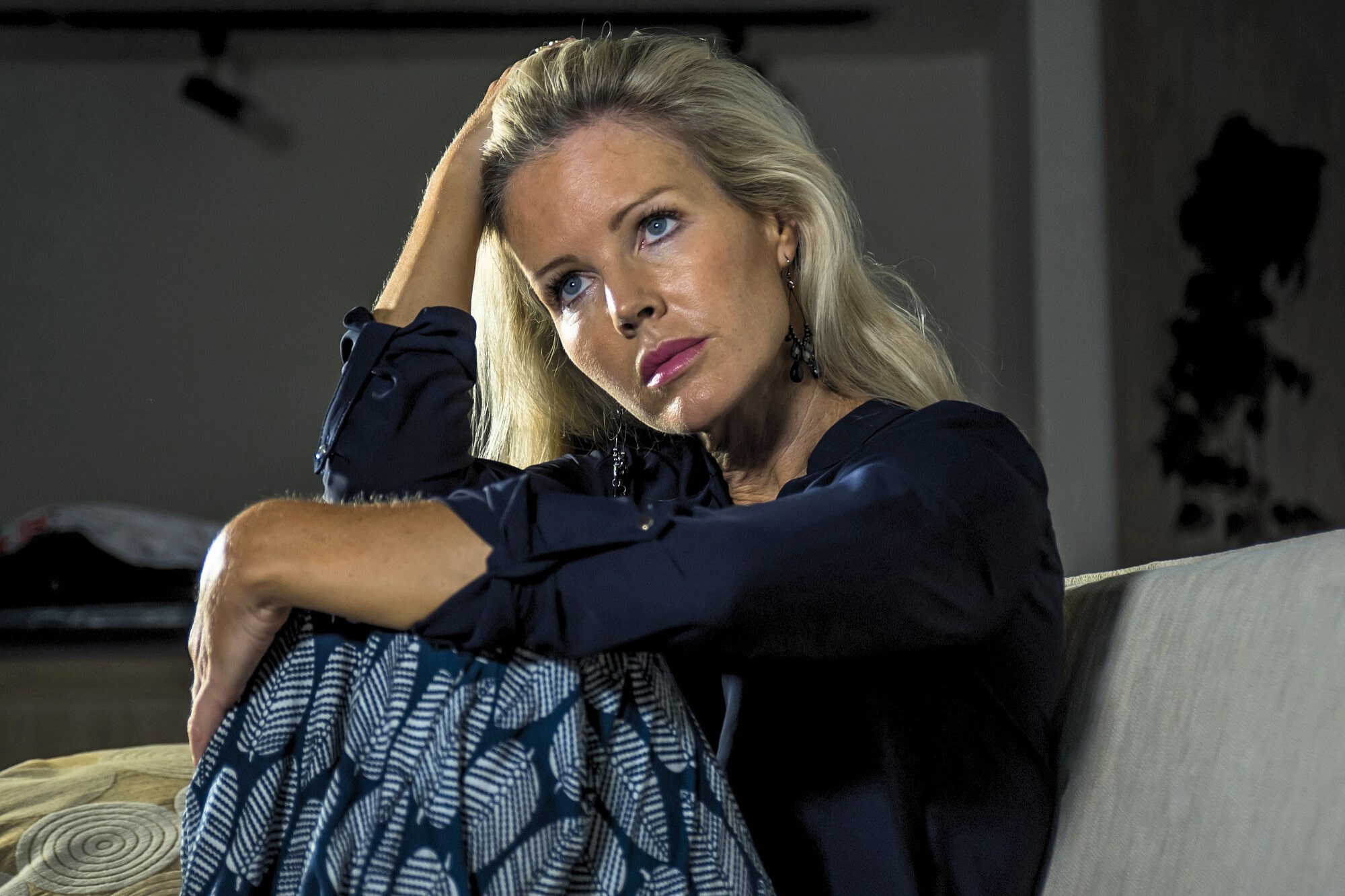 A woman with long blonde hair sits on a couch, resting her arm on her head and looking thoughtfully into the distance. She wears a navy blouse and patterned skirt. The lighting is dim, creating a contemplative mood.